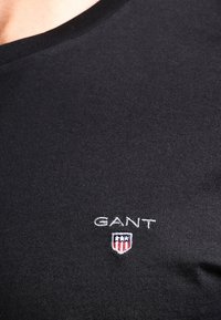 Nærbillede af sort GANT skjorte med hvidt logo og et lille skjoldemblem med stjerner og striber på brystet.