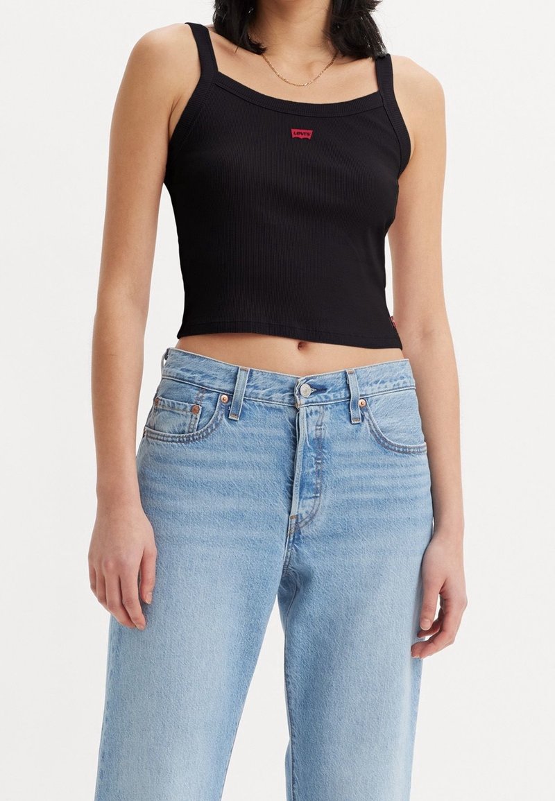 Černý žebrovaný crop top se širokými ramínky a malou červenou etiketou. V kombinaci se světle modrými džínami s vysokým pasem a klasickým knoflíkovým zapínáním.