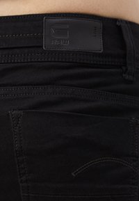 Jean en denim noir avec une finition texturée, présentant un patch logo G-Star RAW à la taille et des coutures détaillées le long des poches.