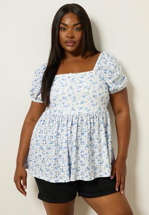 Frau mit langem, glattem Haar, die ein weißes Oberteil mit blauem Blumenmuster und Puffärmeln trägt, kombiniert mit schwarzen Shorts, steht an einer Wand.