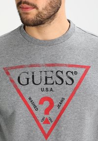 Camiseta de algodón gris con un gran diseño de triángulo rojo que incluye el texto "GUESS U.S.A." y signos de interrogación; mangas cortas y cuello redondo.