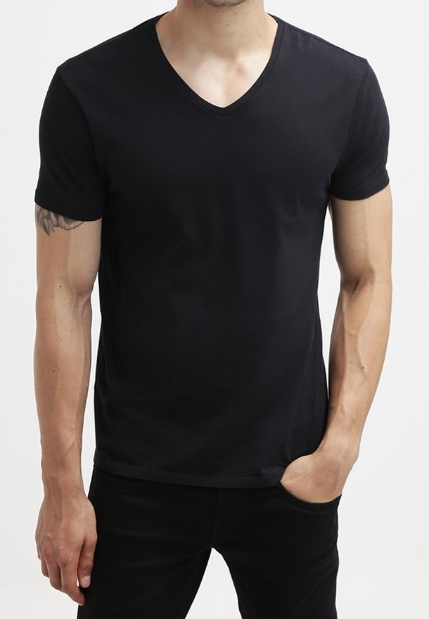 Mand iført en almindelig sort V-hals kortærmet T-shirt med den ene hånd i lommen, som viser en tatovering på venstre overarm, stående foran en hvid baggrund.