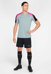 Maglia sportiva Nike grigia chiara con accenti rosa e neri, abbinata a pantaloni corti neri e calze nere al ginocchio, esposta su un manichino.
