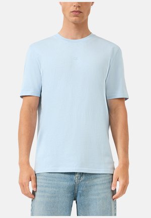 Mężczyzna ubrany w prostą, jasnoniebieską koszulkę z krótkim rękawem i jasnoniebieskie dżinsy, stojący na białym tle.