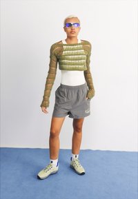 Háčkovaný zelený top s výrezmi, šedé šortky a svetlé topánky. Model má na sebe modré slnečné okuliare, stojí na modrom povrchu proti bielej pozadi.