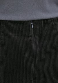 Gros plan sur un pantalon en velours côtelé noir avec des stries verticales visibles et une fermeture éclair à l'avant, sous un haut en maille gris.