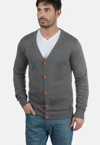 Grå strikket cardigan med V-hals, med treskinnknapper foran og ribbestrikkede mansjetter. Kombinert med en hvit T-skjorte og blå jeans.