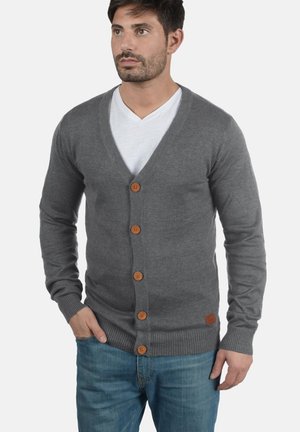 Grå strikket cardigan med V-hals, med treskinnknapper foran og ribbestrikkede mansjetter. Kombinert med en hvit T-skjorte og blå jeans.