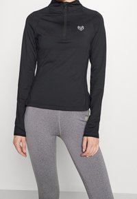 Černý dlouhý rukáv atletického topu s čtvrtinovým zipem a geometrickým srdíčkovým logem, spárovaný se šedými úzkými legínami. Hladký povrch látky.