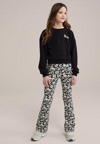 Fekete, rövidített kapucnis pulóver "Le Magis" felirattal, magas derekú, bővülő leggingssel párosítva, amely fehér virágmintával díszített. Szürke sportcipő.