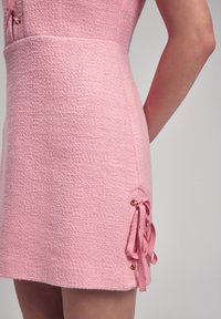 Rosa strukturiertes Kleid mit einer taillierten Silhouette, das eine seitliche Schleifenbindung und metallische Ösen am Saum aufweist.