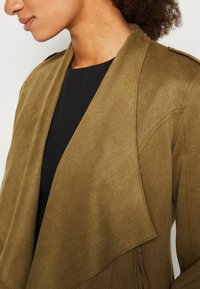 Gros plan d'une personne portant une veste en daim marron avec de larges revers sur un top noir, montrant l'épaule gauche et la zone du cou.