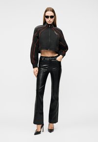 Γυναίκα που φοράει μαύρο cropped μπουφάν με πορτοκαλί περιθώριο, μαύρο υφασμάτινο παντελόνι, μαύρες μυτερές γόβες και μαύρα γυαλιά ηλίου, στέκεται με το ένα χέρι στην τσέπη.