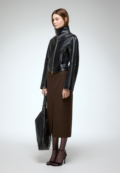 Chaqueta de cuero negra con cuello alto y costuras asimétricas. Falda de lana marrón con una abertura frontal. Sandalias negras de tacón alto y un bolso grande.