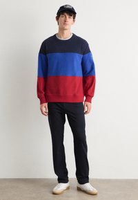 Color-blocked sweatshirt featuring navy, blue, and red sections. Cotton blend, crew neck, relaxed fit, worn with black pants and white sneakers.