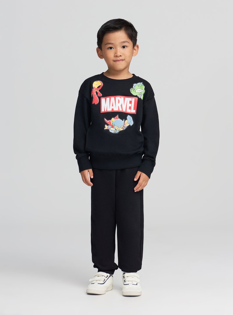 Ragazzo giovane con felpa nera Marvel con personaggi dei cartoni animati, pantaloni neri e scarpe da ginnastica bianche, in piedi davanti a uno sfondo grigio uniforme.