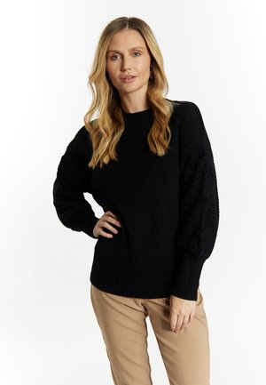 Czarny sweter robiony na drutach z prążkowanym dołem i teksturowanymi rękawami w kształcie balonu, w połączeniu z beżowymi spodniami. Minimalistyczny design, gładka faktura materiału.