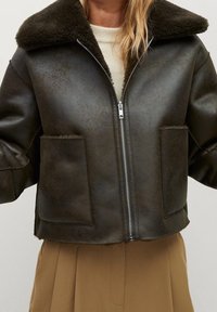 Veste en cuir marron foncé avec grand col en shearling, fermeture éclair devant et deux poches avant, portée sur un haut blanc et un pantalon beige.