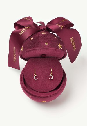 Petites boucles d'oreilles en forme de croissant de lune en or avec diamants présentées dans un écrin rond en velours bordeaux décoré d'étoiles dorées et d'un nœud assorti.