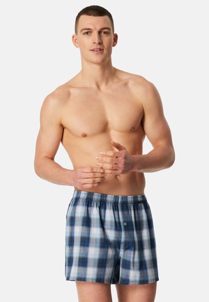 Young man with short brown hair, shirtless, wearing blue and white plaid boxer shorts, standing against a plain light background.