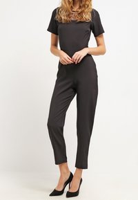 Svart byxdress med korta ärmar, figurnära design och byxor som når anklarna. Slät tyg med en strukturerad detalj vid kragen. Bärs med svarta klackar.