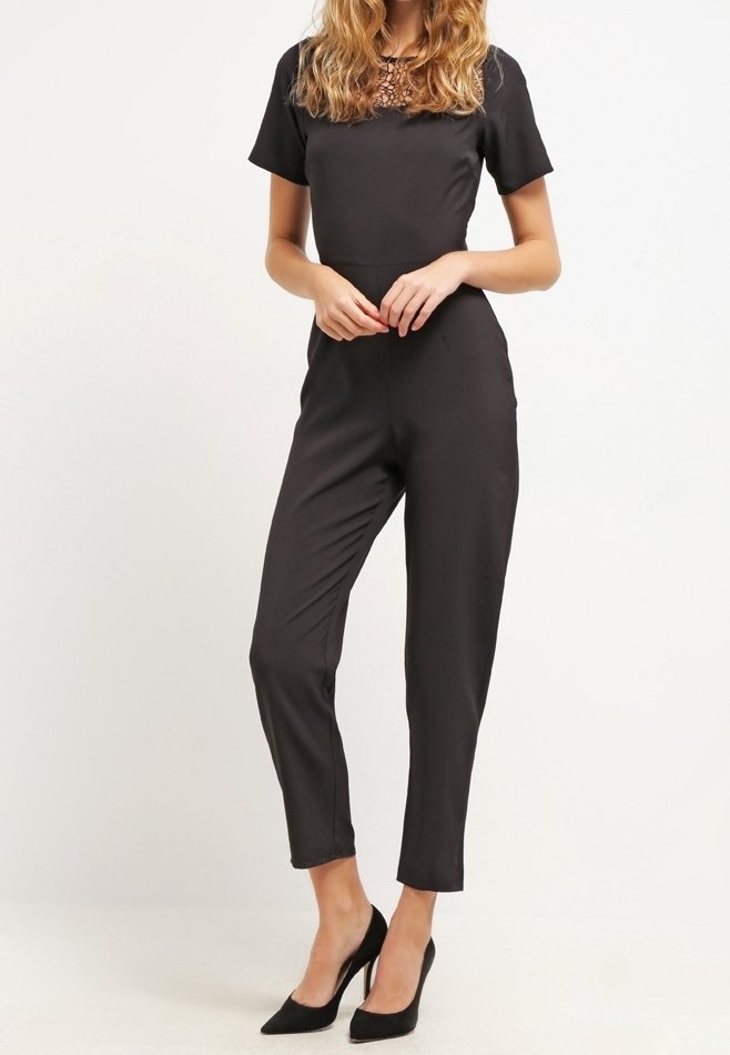 Svart byxdress med korta ärmar, figurnära design och byxor som når anklarna. Slät tyg med en strukturerad detalj vid kragen. Bärs med svarta klackar.
