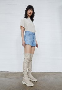 Beige læder støvler op til låret med tekstureret overflade og hvide gummisåler, parret med lyse denim shorts og en beige skjorte med knapper.