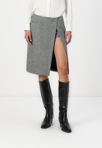 Saia envolvente em mistura de lã cinzenta com uma fenda alta, combinada com botas de couro brilhante até ao joelho pretas. Apresenta bolsos e um design ajustado.