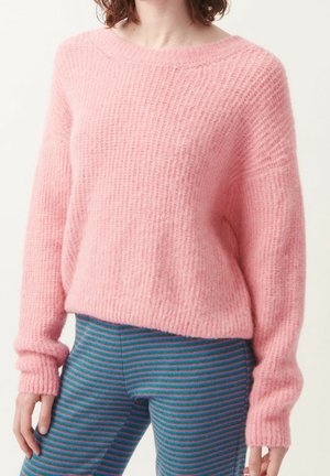 Mujer con un suéter rosa suave y texturizado y pantalones a rayas azul verdoso, de pie con los brazos relajados a los lados.