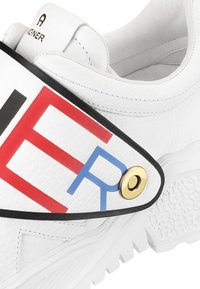 Baskets blanches en cuir texturé, avec une large bride arborant des lettres rouges, noires et bleues, une quincaillerie dorée et une semelle blanche robuste.