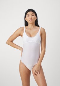 Hvit one-piece badedrakt med tynne stropper, v-hals og et nettingpanel foran. Stoffet er glatt og lett gjennomsiktig.