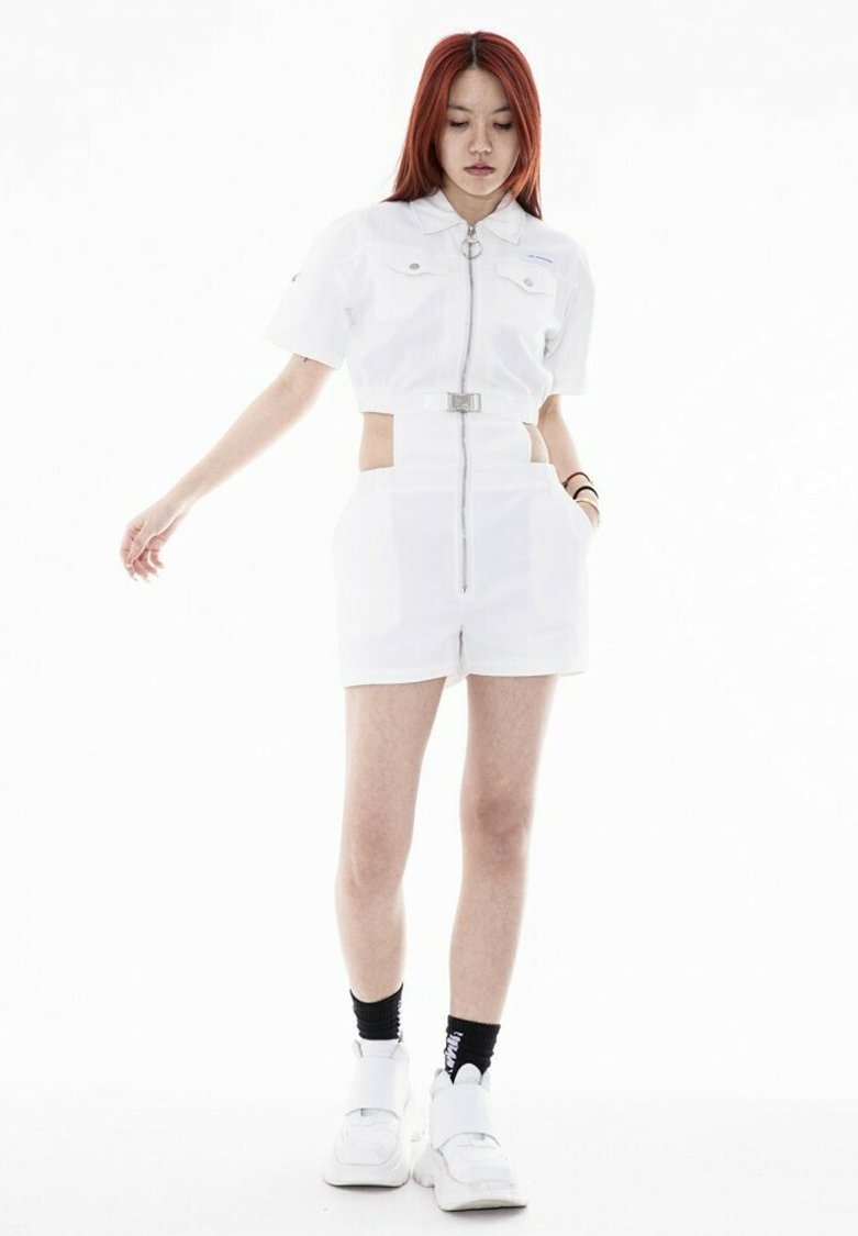 Camisa blanca corta con cremallera frontal y dos bolsillos en el pecho, combinada con pantalones cortos de cintura alta que tienen recortes laterales y un detalle de cinturón.