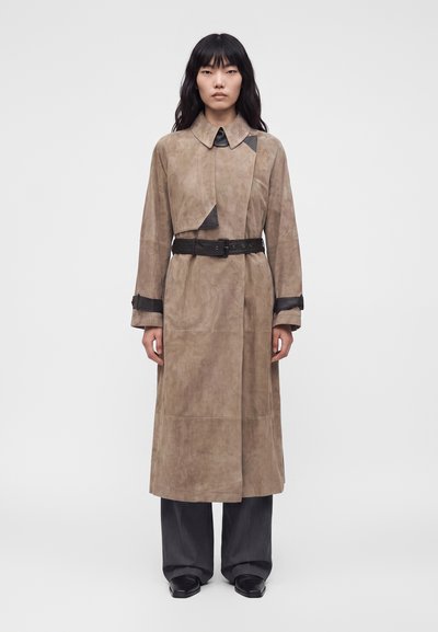 Femme portant un long trench-coat taupe avec des accents en cuir noir et une ceinture, associé à un pantalon large foncé et des chaussures noires, debout sur fond blanc.