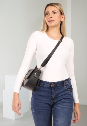 Blonde woman wearing white long-sleeve top and blue jeans, carrying black crossbody bag with Italian flag charm, standing indoors.