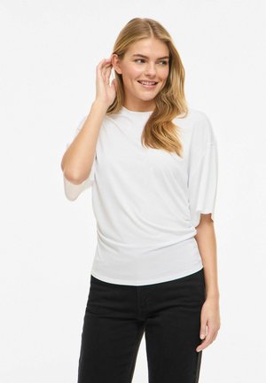 Ung kvinne med langt blondt hår som har på seg en hvit kortermet skjorte og svarte bukser, smilende og berører øret sitt.