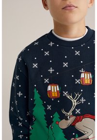 Marinblå sweatshirt med vita snöflingemönster, tecknad ren med en röd halsduk och skidgondoler bland gröna träd.