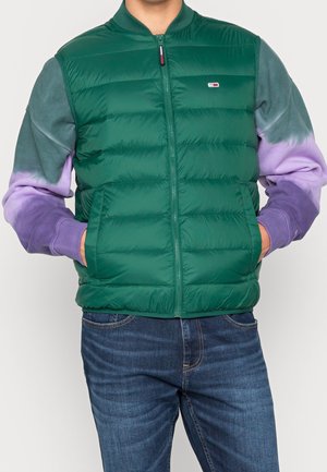 Homme portant une veste matelassée verte sur une chemise à manches longues dégradée gris et violet, les mains dans les poches de la veste, associée à un jean bleu.