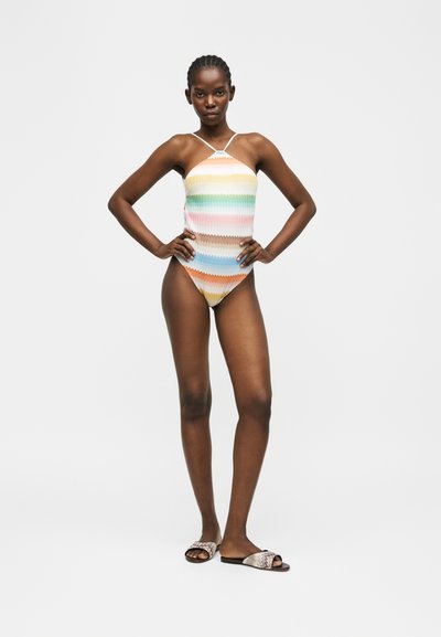 Γυναίκα που στέκεται με τα χέρια στη μέση, φοράει ολόσωμο μαγιό με λουράκι στον λαιμό και πολύχρωμες ριγέ ρίγες, καθώς και σχέδια σανδάλια πάνω σε λευκό φόντο.