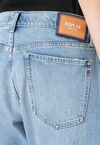 Világoskék farmernadrág barna bőr címkével, amelyen az áll: "REPLAY SINCE 1981." Jellemzője egy hátsó zseb díszvarrással.