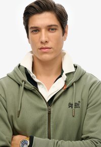 Sudadera verde oliva con cremallera, con bolsillos frontales, capucha ajustable y logo; superpuesta a una camisa blanca con cuello y ribete marino.