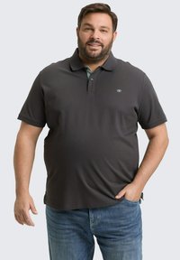 Mężczyzna z brodą, ubrany w ciemnoszary polo z krótkim rękawem i niebieskie dżinsy, stojący z jedną ręką w kieszeni na jednolitym tle.