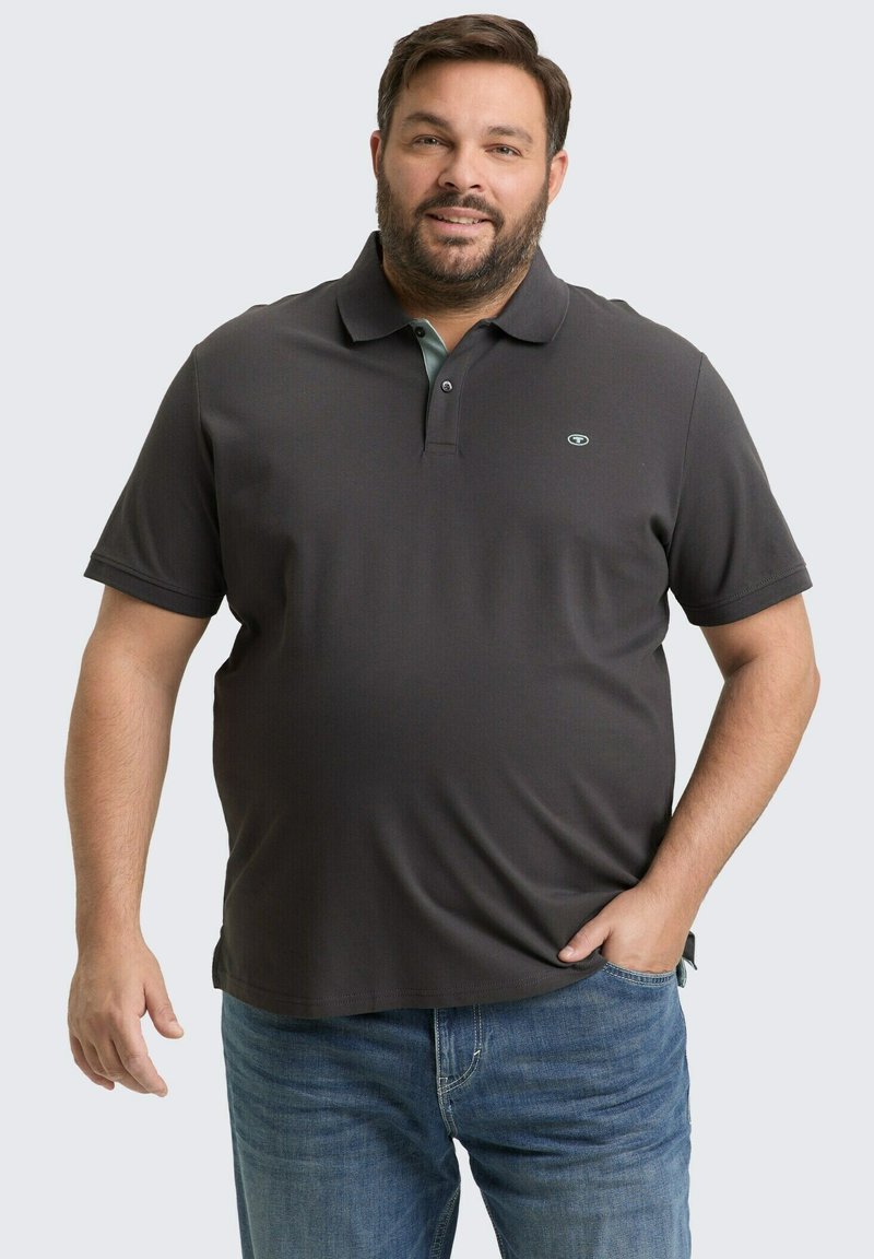 Mężczyzna z brodą, ubrany w ciemnoszary polo z krótkim rękawem i niebieskie dżinsy, stojący z jedną ręką w kieszeni na jednolitym tle.