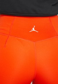 Leggings arancioni con una texture liscia, caratterizzati da cuciture bianche e un logo Jumpman centrato sul retro, abbinati a una maglietta nera.