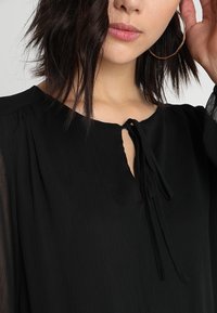 Schwarze Bluse aus leichtem, strukturiertem Stoff. Hat einen Schlüssel-Ausschnitt mit einer gebundenen Detaillierung und langen, leicht durchsichtigen Ärmeln.