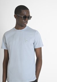 T-shirt in cotone azzurro chiaro con scollo tondo, maniche corte e una piccola etichetta con logo sul petto. Modello che indossa occhiali da sole neri.