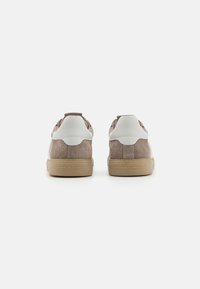 Une paire de baskets en daim beige avec des accents blancs au niveau du talon et des semelles en gomme, vues de derrière sur un fond blanc uni.
