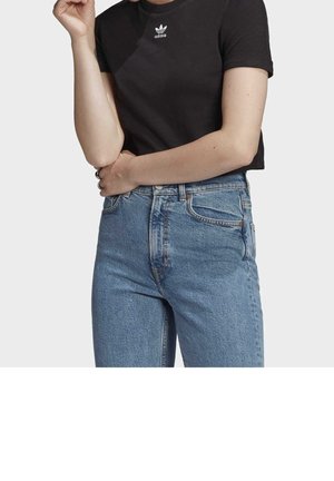 Persoon die een zwart Adidas t-shirt en lichtblauwe high-waisted jeans draagt, staat met één arm gekruist en de andere opgetild bij het gezicht.