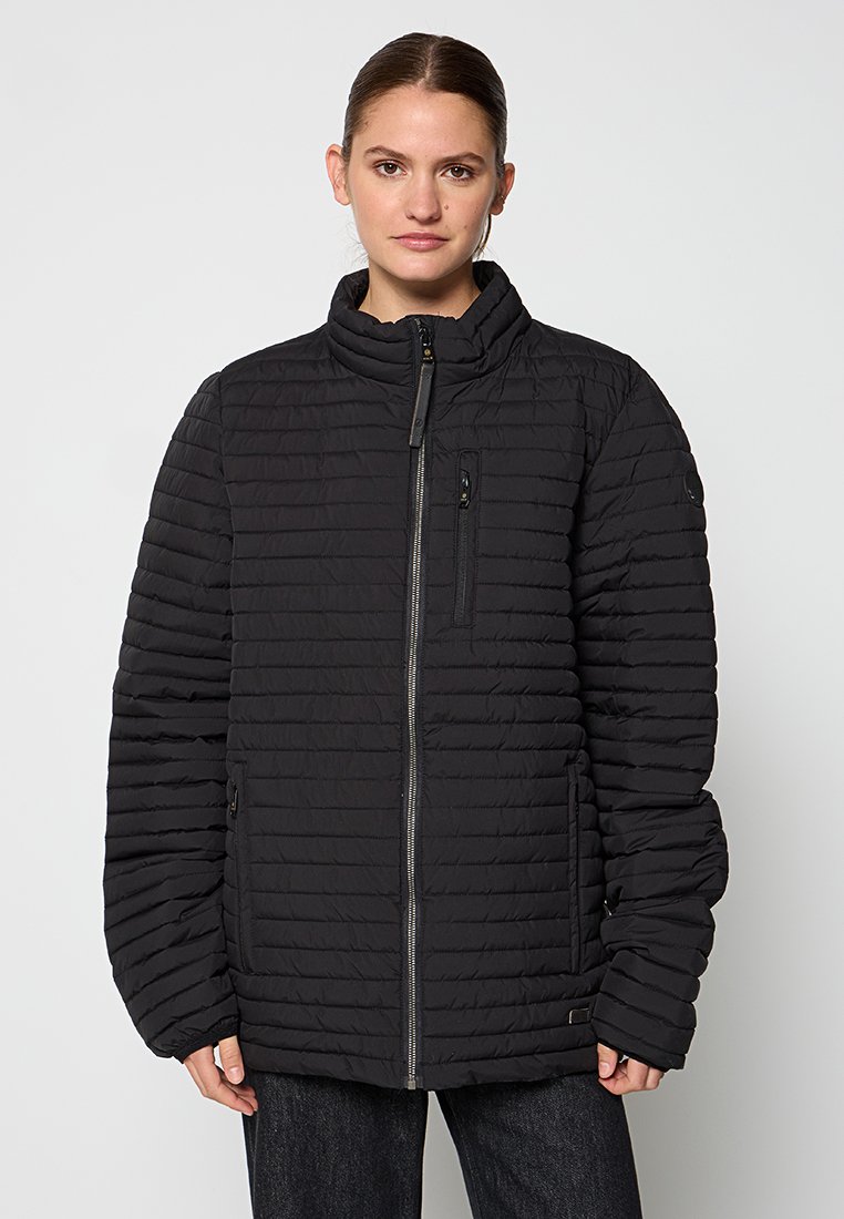 Femme portant une veste zippée matelassée noire avec un col haut, une poche poitrine à l'avant et des poches latérales, se tenant devant un fond uni.