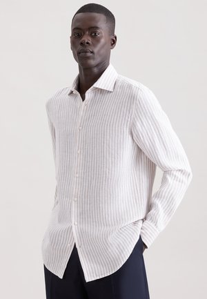 Homme portant une chemise à manches longues beige clair à rayures blanches et un pantalon noir, debout avec une main dans la poche contre un fond uni.