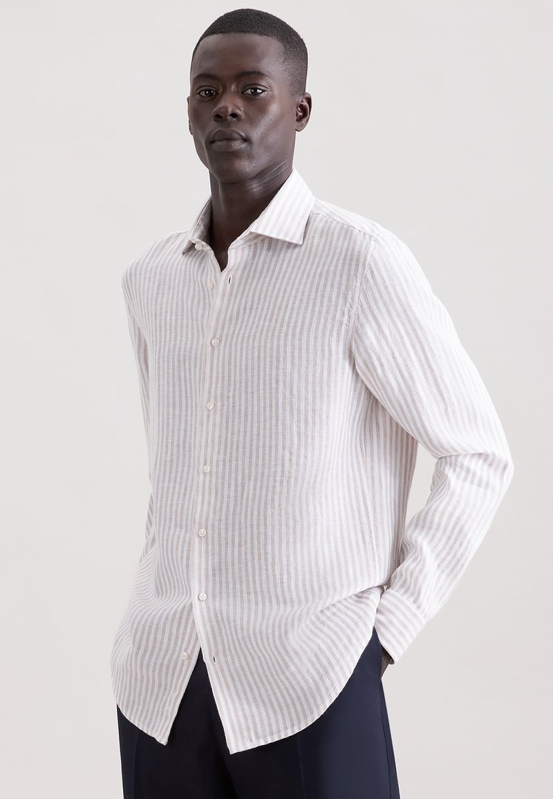 Homme portant une chemise à manches longues beige clair à rayures blanches et un pantalon noir, debout avec une main dans la poche contre un fond uni.